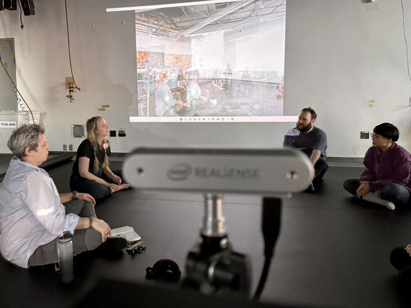 Un groupe de personnes est assis en cercle sur le sol d’une salle, face à un grand écran projetant une captation par caméra Intel RealSense, fixée sur un trépied au premier plan et orientée vers le groupe.