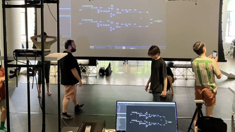 L’image montre une salle avec un grand écran projetant une interface de programmation visuelle composée de blocs et de connexions. Au premier plan, un ordinateur affiche la même interface, posé sur un support avec du matériel technique autour. Plusieurs personnes se déplacent ou observent dans la salle.