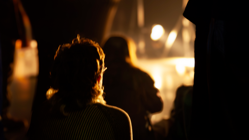 Plan rapproché du profil d’une personne assise, éclairée par une lumière orange, dans une foule regardant une performance.